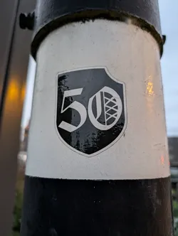 A black and white sticker featuring the number 50 inside a shield shape. The number is in an old-style font and includes a smaller graphic within the shield.