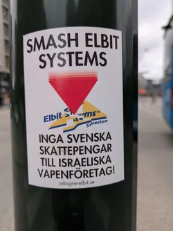 The sticker, adhered to a dark pole, prominently displays text against Elbit Systems. The top reads "SMASH ELBIT SYSTEMS" in bold black letters. Below this, a red inverted triangle symbol is integrated into a modified logo that mimics Elbit Systems' branding, with the legible text "Elbit S-stems™ Sweden". The bottom part of the sticker carries a message in Swedish: "INGA SVENSKA SKATTEPENGAR TILL ISRAELISKA VAPENFÖRETAG!", which translates to "NO SWEDISH TAX MONEY TO ISRAELI WEAPONS COMPANIES!". At the very bottom, the website "stängnerelbit.se" is printed, further emphasizing the call to action against Elbit.