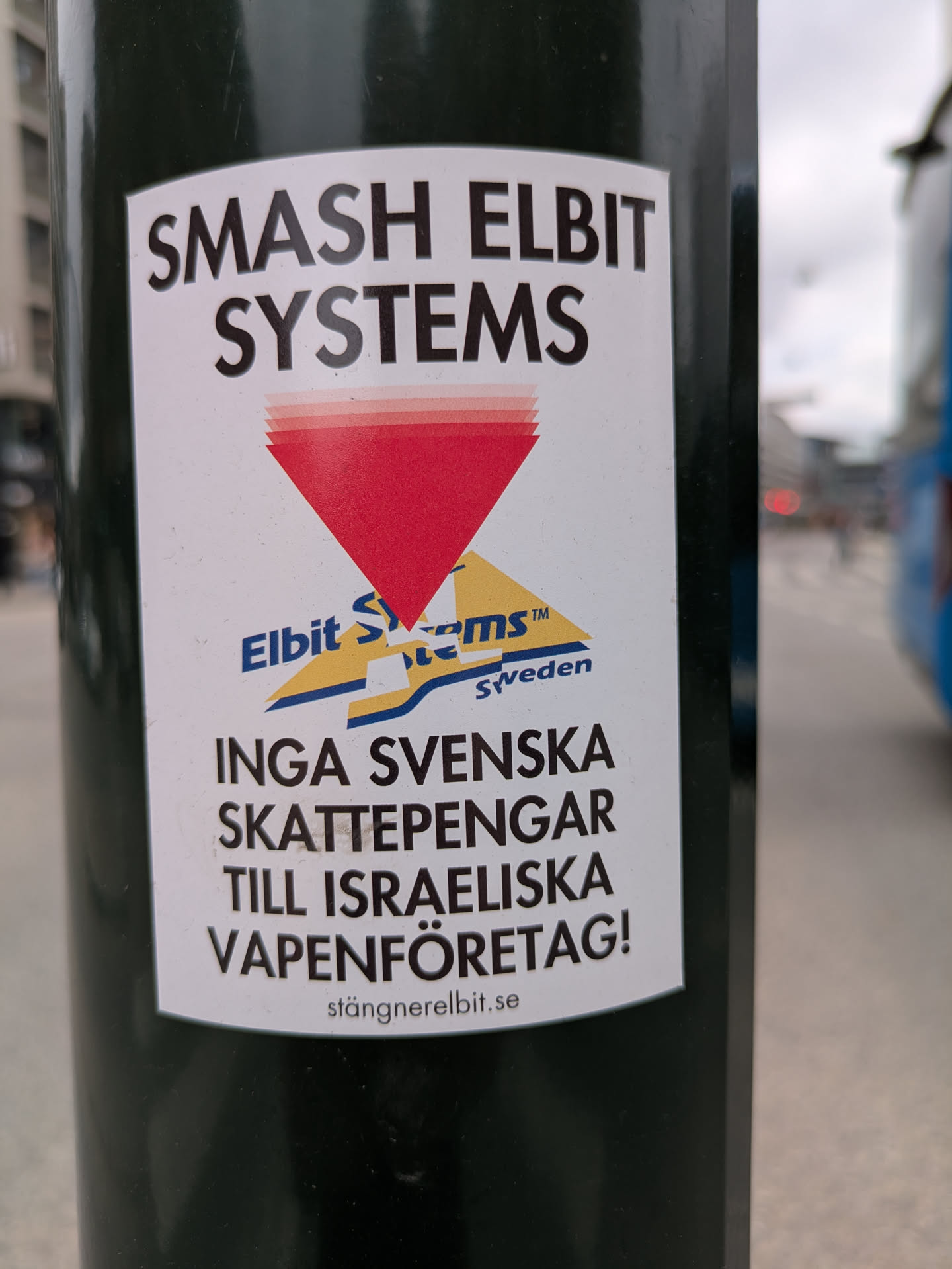 The sticker, adhered to a dark pole, prominently displays text against Elbit Systems. The top reads "SMASH ELBIT SYSTEMS" in bold black letters. Below this, a red inverted triangle symbol is integrated into a modified logo that mimics Elbit Systems' branding, with the legible text "Elbit S-stems™ Sweden". The bottom part of the sticker carries a message in Swedish: "INGA SVENSKA SKATTEPENGAR TILL ISRAELISKA VAPENFÖRETAG!", which translates to "NO SWEDISH TAX MONEY TO ISRAELI WEAPONS COMPANIES!". At the very bottom, the website "stängnerelbit.se" is printed, further emphasizing the call to action against Elbit.