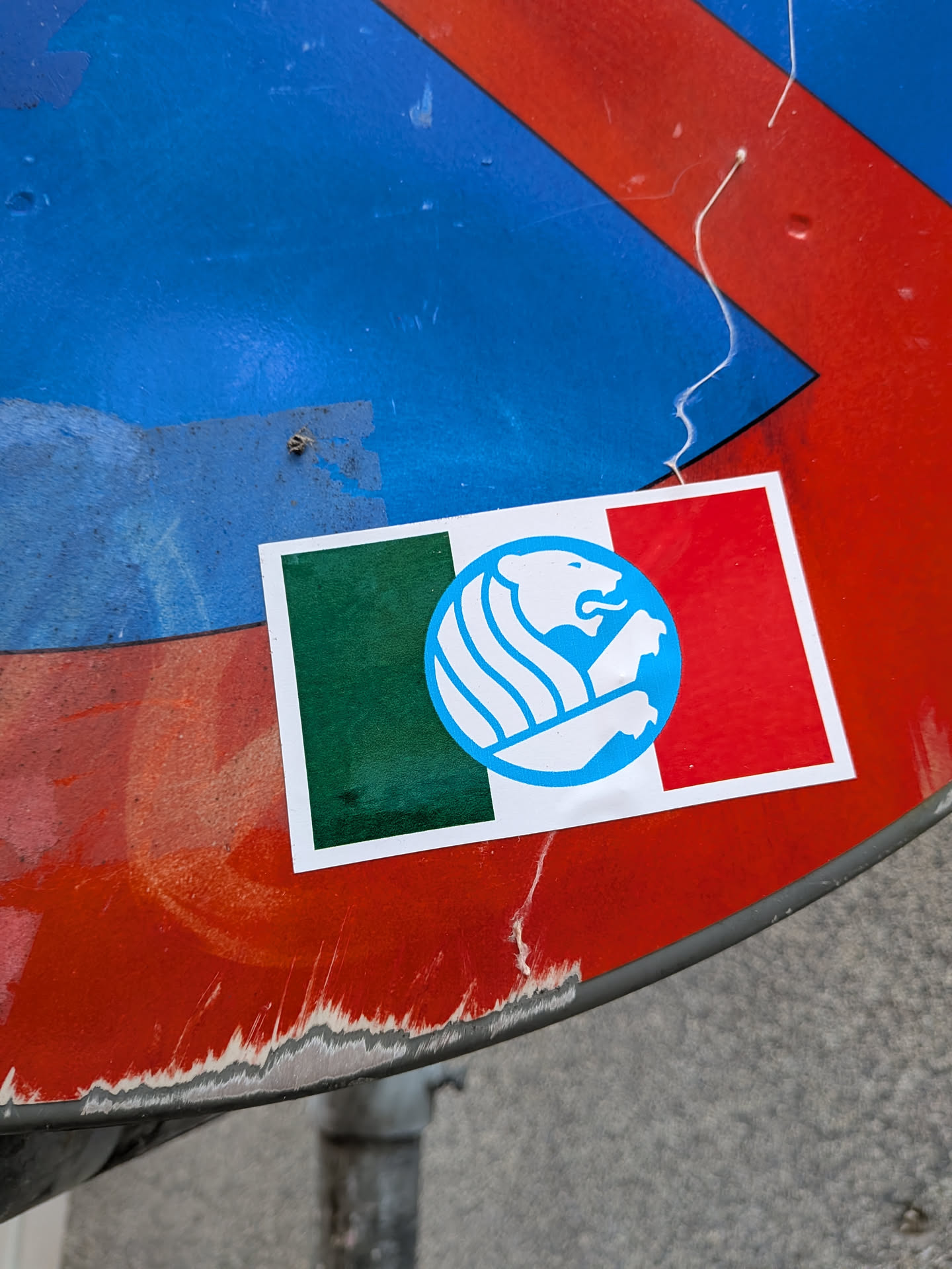 A rectangular sticker with a thin white border depicting the Italian tricolor (green, white, and red vertical bands). Centered over the white stripe is a light blue circular medallion containing a minimalist, stylized silhouette of a heraldic lion in white, depicted facing right with its mouth open and forepaws raised.