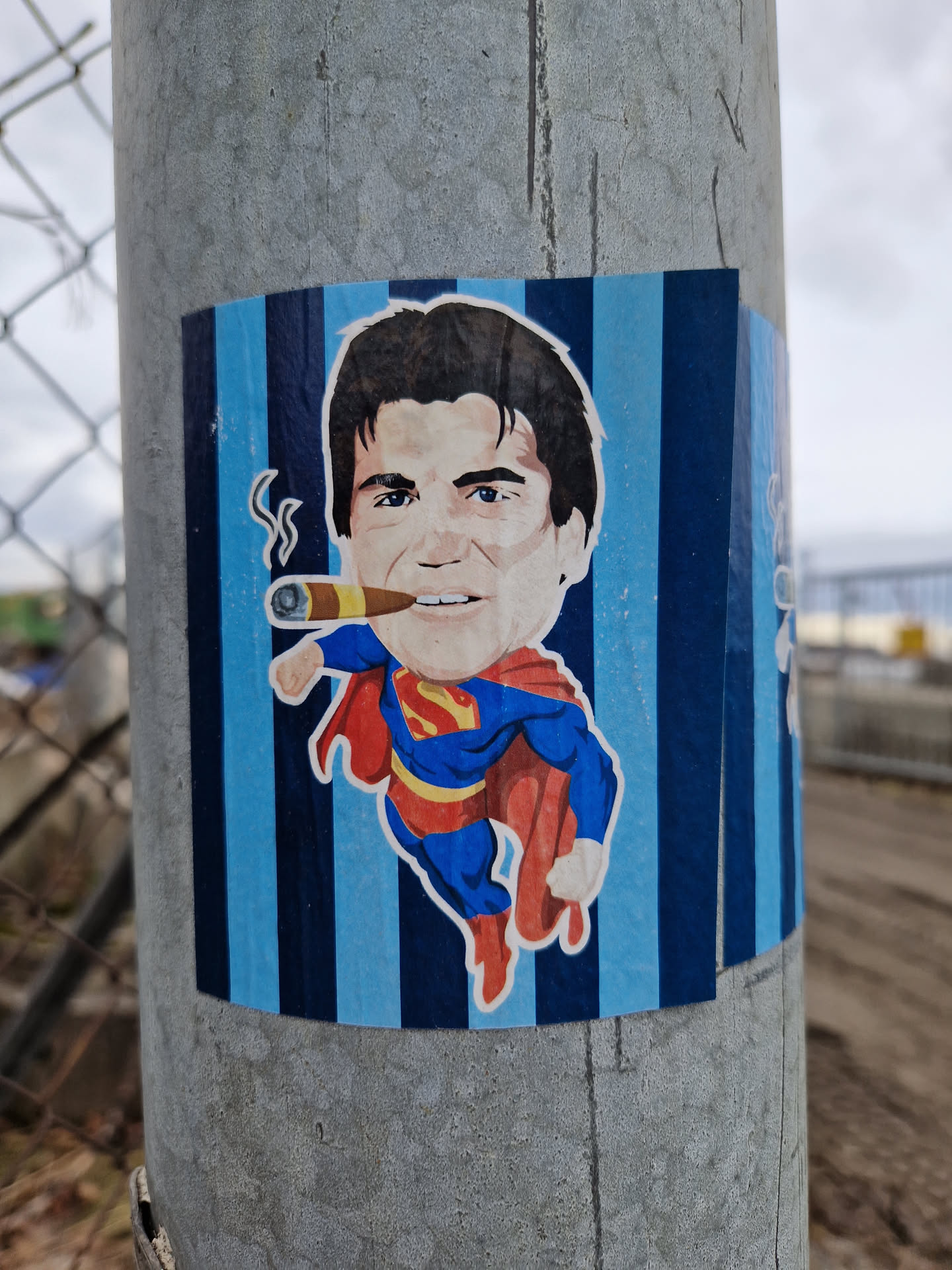 A sticker featuring a caricature of Superman smoking a cigar against a blue and black striped background.