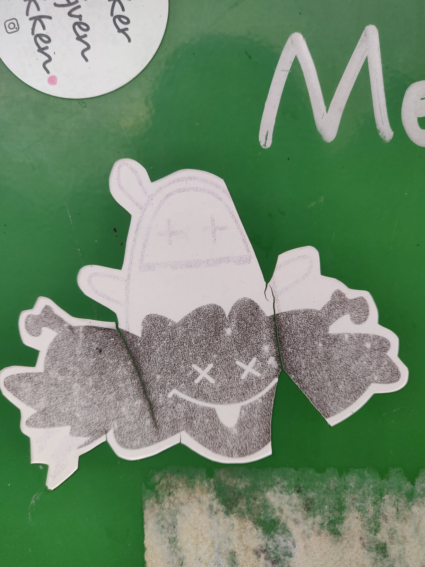 This sticker features a dark-grey monster with a cute, spooky design. It seems to be made of a textured material. The monster has X-shaped eyes and small fangs.  A lighter-colored, possibly white, ghost-like figure is partially visible above it. The sticker is adhered to a green surface.