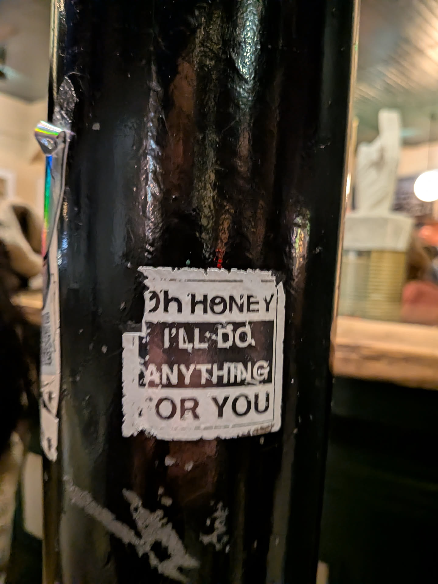 The sticker is rectangular with a distressed white background and bold black text. The text reads 'Oh HONEY I'LL DO ANYTHING FOR YOU' arranged in three lines. The edges of the white background are ragged and peeling, giving it a worn, grungy appearance. The sticker is affixed to a dark, possibly black, textured metal pole.