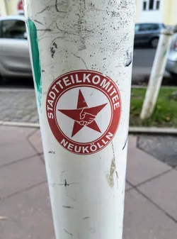 A red and white sticker with a red star and clasped hands in the center. The text around the star says "Stadtteilkomitee Neukölln".