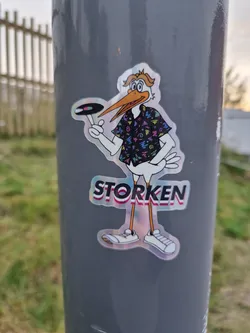 A holographic sticker of a cartoon stork wearing a retro-style shirt and holding a vinyl record. The stork is named "Storken". The sticker has an iridescent finish and appears to be a promotional item.