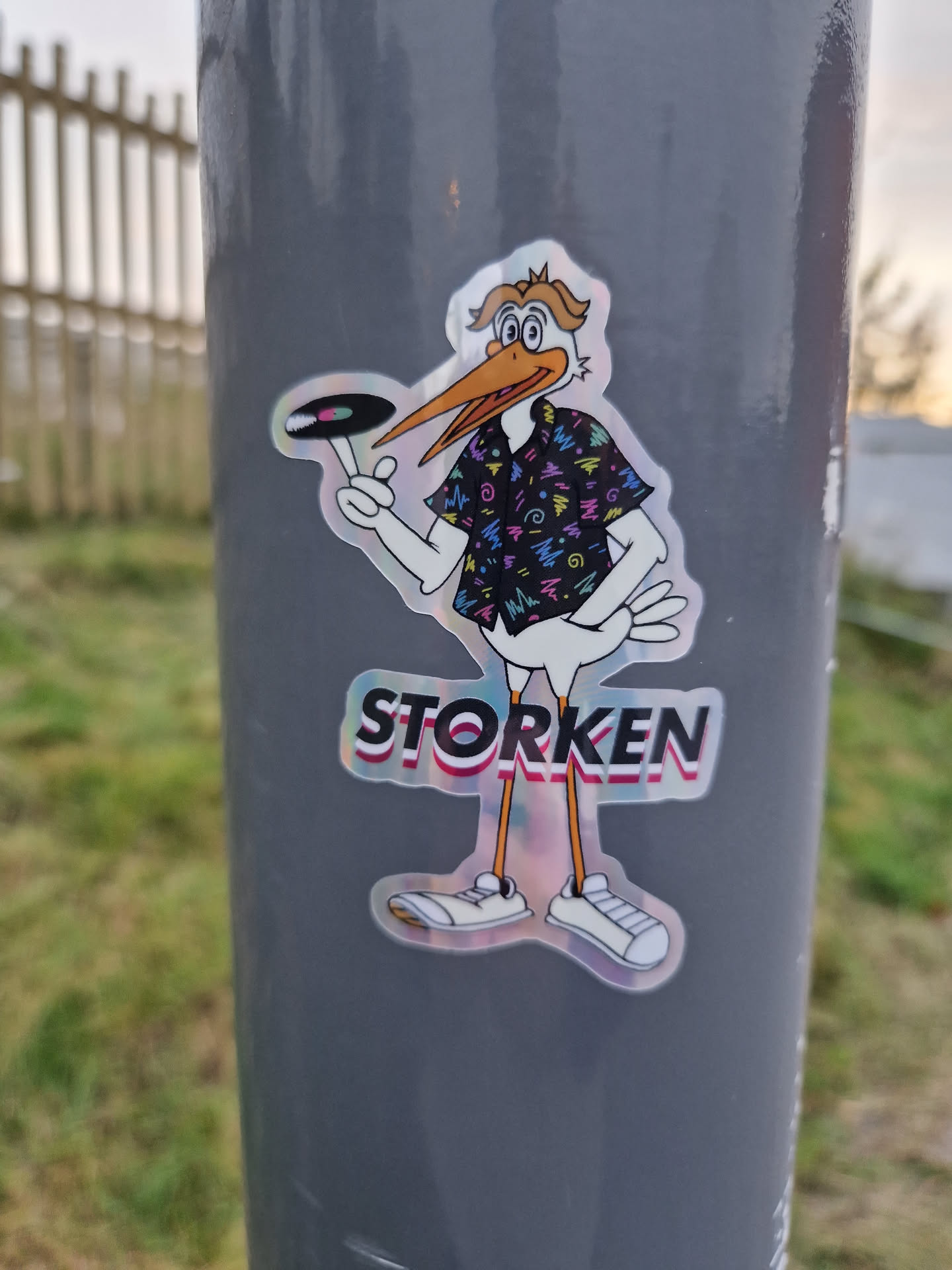 A holographic sticker of a cartoon stork wearing a retro-style shirt and holding a vinyl record. The stork is named "Storken". The sticker has an iridescent finish and appears to be a promotional item.