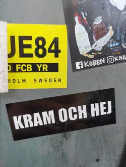 A black sticker with white text that says "Kram och hej" which means "Hug and hi" in Swedish. It's attached to a gray surface with other stickers around it.