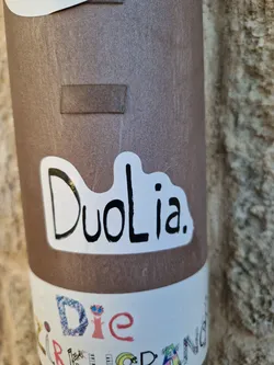 A simple white sticker with the word DuoLia. written in black. The sticker has a slightly irregular, cloud-like shape. It's affixed to a brown cylindrical object, which appears to be a post or a similar structure. There's another sticker partially visible below DuoLia sticker.