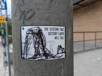 The sticker is a rectangular, black and white image applied to a textured concrete pole. Its main visual element is a large, intricately drawn high-voltage power line tower (pylon) that appears to be collapsing or already fallen, its metal structure splayed across the ground. In the background, other pylons are visible, some standing, creating a sense of a landscape of industrial decay. The drawing style is stark, using thick black lines on a white background. To the right of the image, bold, capitalized text is prominently displayed. In the top-left corner, there is a small circular logo with a green background and a white letter 'A' inside.