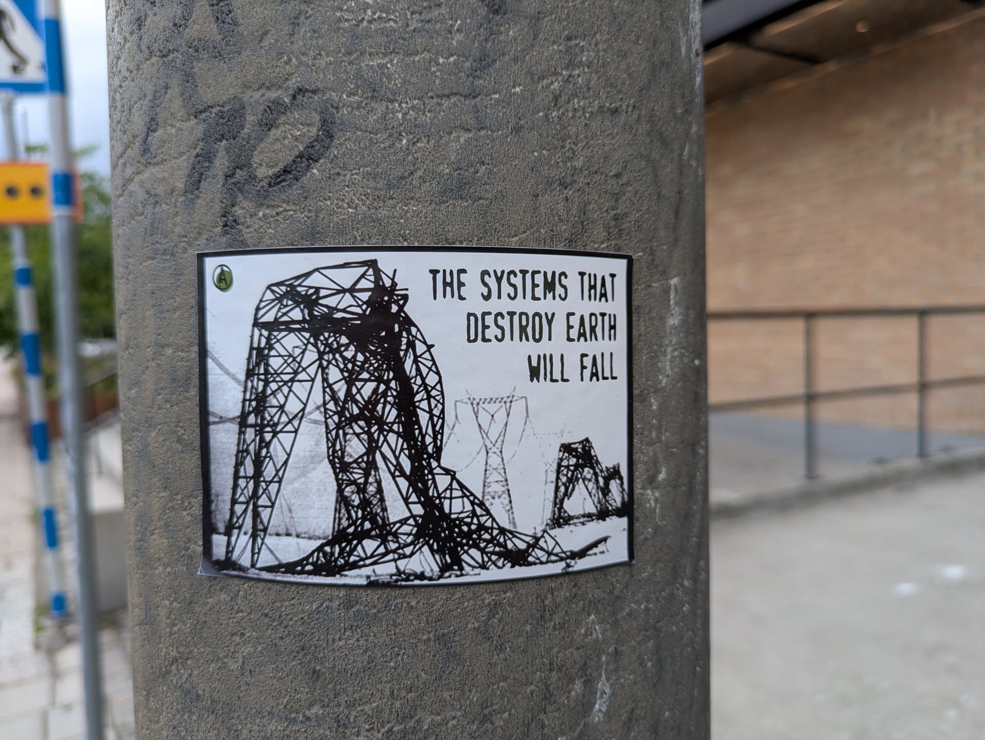The sticker is a rectangular, black and white image applied to a textured concrete pole. Its main visual element is a large, intricately drawn high-voltage power line tower (pylon) that appears to be collapsing or already fallen, its metal structure splayed across the ground. In the background, other pylons are visible, some standing, creating a sense of a landscape of industrial decay. The drawing style is stark, using thick black lines on a white background. To the right of the image, bold, capitalized text is prominently displayed. In the top-left corner, there is a small circular logo with a green background and a white letter 'A' inside.