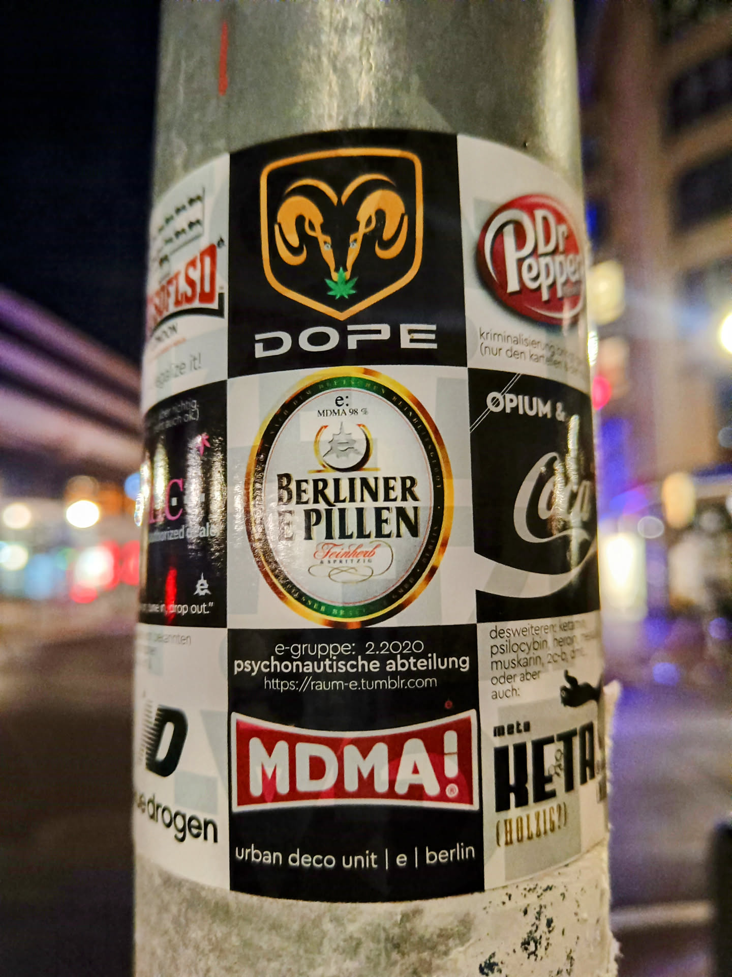 A collage-style sticker featuring various elements, including a prominent Ram head logo with a cannabis leaf in its mouth above the word 'DOPE', a Berliner Weisse beer label, and a large red 'MDMA!' declaration. There is additional text indicating the group e-gruppe and the date 2.2020, along with a Tumblr URL: https://raum-e.tumblr.com. The bottom displays the text "urban deco unit | e | berlin". The sticker is affixed to a metal pole alongside other stickers.