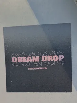 Black sticker with the text DREAM DROP in pink letters.  The text is surrounded by a graphic design. A website address is visible at the bottom. 