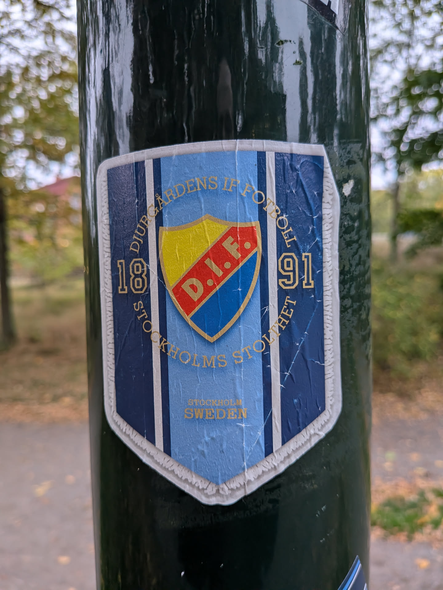 A shield-shaped sticker featuring the Djurgårdens IF Fotboll emblem adhered to a dark green pole. The background consists of vertical stripes in navy blue, light blue, and thin white lines. In the center sits the club's shield crest divided into yellow, red, and blue sections, displaying the initials 'D.I.F.' on a diagonal red sash. Surrounding the inner crest are gold-toned inscriptions reading 'DJURGÅRDENS IF FOTBOLL' along the top curve, 'STOCKHOLMS STOLTHET' along the bottom curve, and the founding year '18' and '91' on either side. Near the pointed base, 'STOCKHOLM SWEDEN' is printed in gold lettering. The sticker is bordered in white and exhibits visible crinkling and weathering.