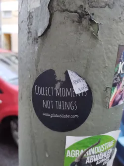 A round black and white sticker on a grey pole. The text on the sticker says "Collect Moments Not Things" and a website address: www.globustliebe.com