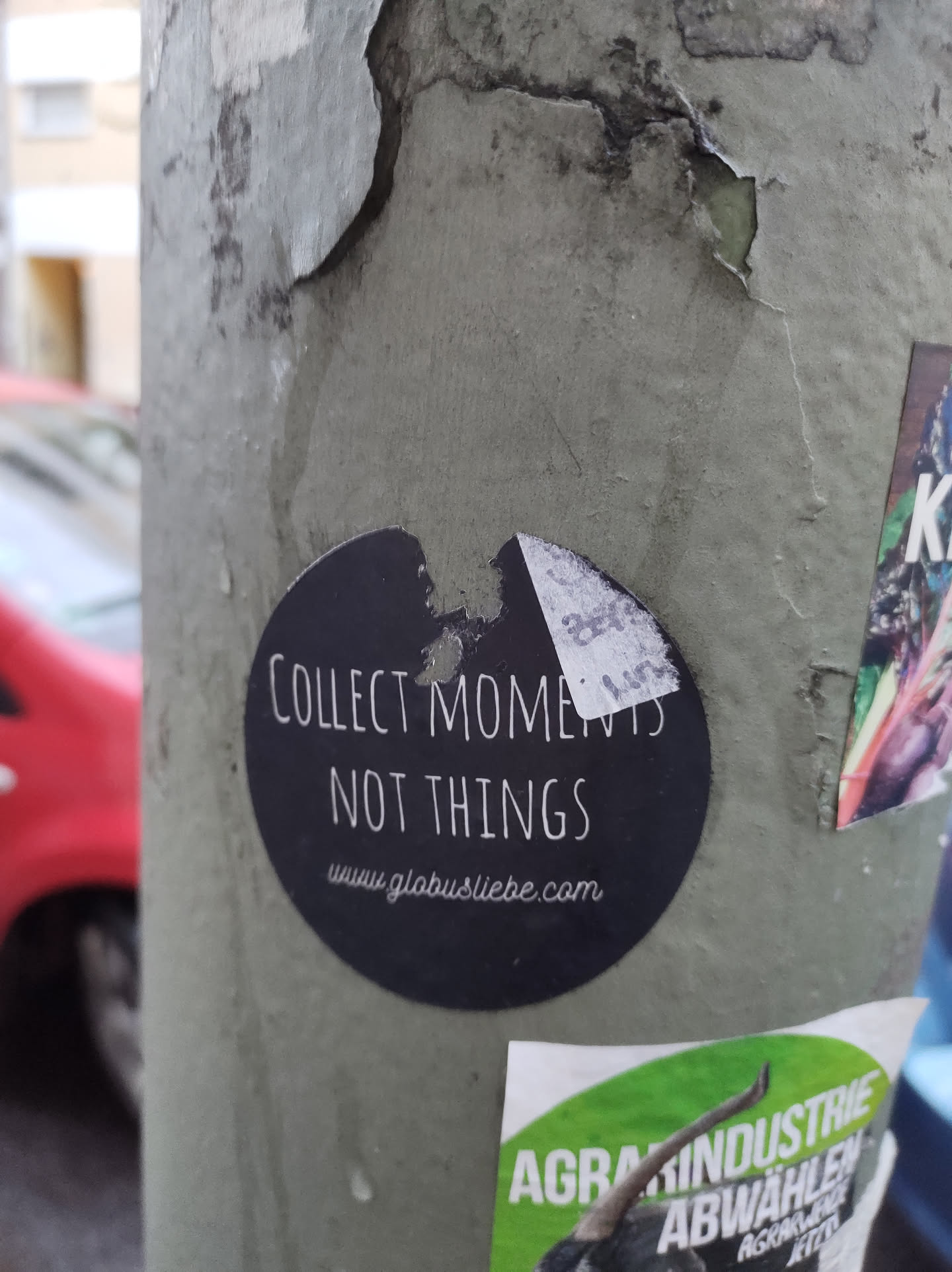 A round black and white sticker on a grey pole. The text on the sticker says "Collect Moments Not Things" and a website address: www.globustliebe.com