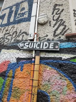 A black sign with the word SUICIDE written in white paint is attached to a pole on a wall covered in graffiti. The graffiti includes various colors and styles of lettering and abstract designs.  There's a small, almost unnoticeable, sticker on the sign's bottom right corner.