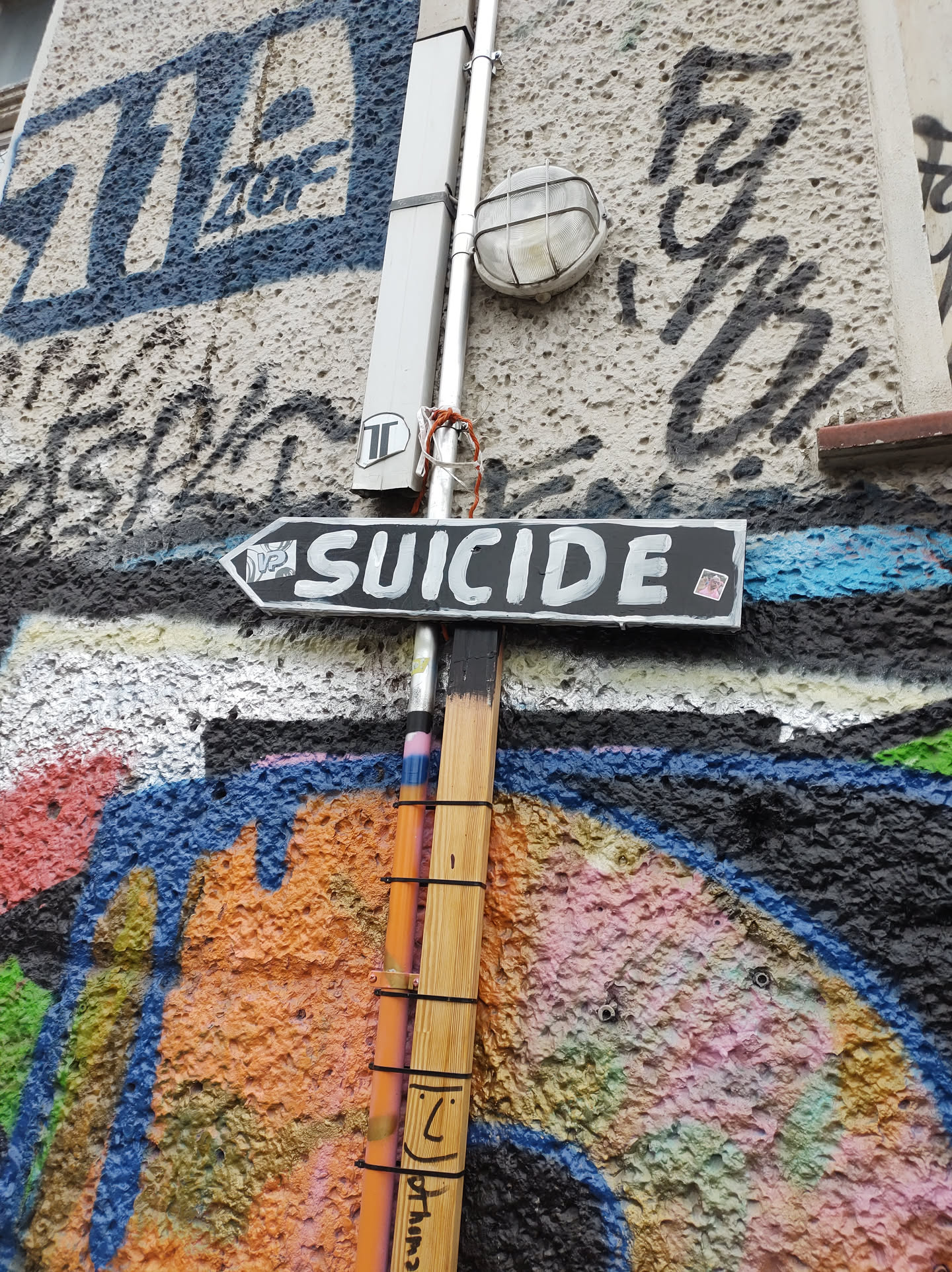 A black sign with the word SUICIDE written in white paint is attached to a pole on a wall covered in graffiti. The graffiti includes various colors and styles of lettering and abstract designs.  There's a small, almost unnoticeable, sticker on the sign's bottom right corner.
