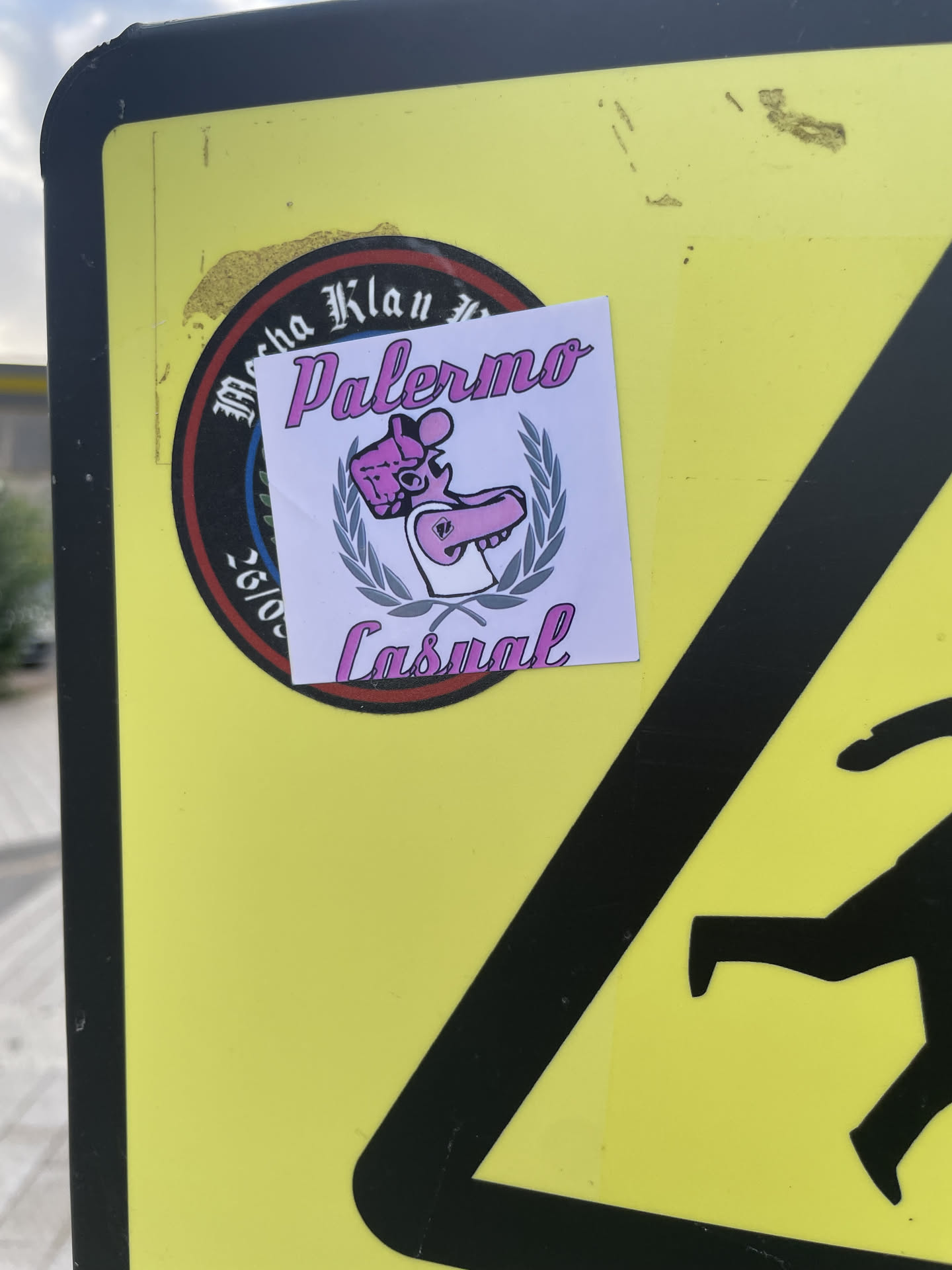 A square white sticker featuring the text 'Palermo' on top and 'Casual' on the bottom in pink script font with black outlines. Centered between the text is a pink cartoon figure wearing a flat cap and a white tank top, enclosed within a greyish-blue laurel wreath. The sticker is pasted over a round black-and-red sticker on a yellow metal sign.