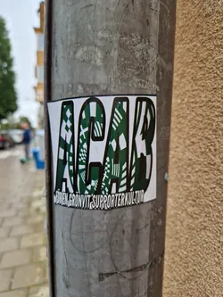 A close-up shot of a sticker featuring the acronym ACAB (All Cops Are Bastards) in bold, stylized lettering. The letters are filled with a geometric, almost camouflage-like pattern in shades of green and white. Below the main text is smaller text that reads  "FöREN, GRÖNVIT, SUPPORTERKULTUR". The sticker is affixed to a weathered gray pole.