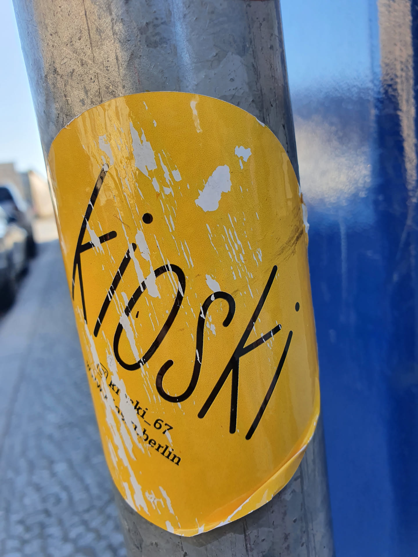 A yellow sticker with the word Kiosk written in black.  The sticker is slightly damaged and worn. Small text at the bottom reads 'ki_ki_67 berlin'.