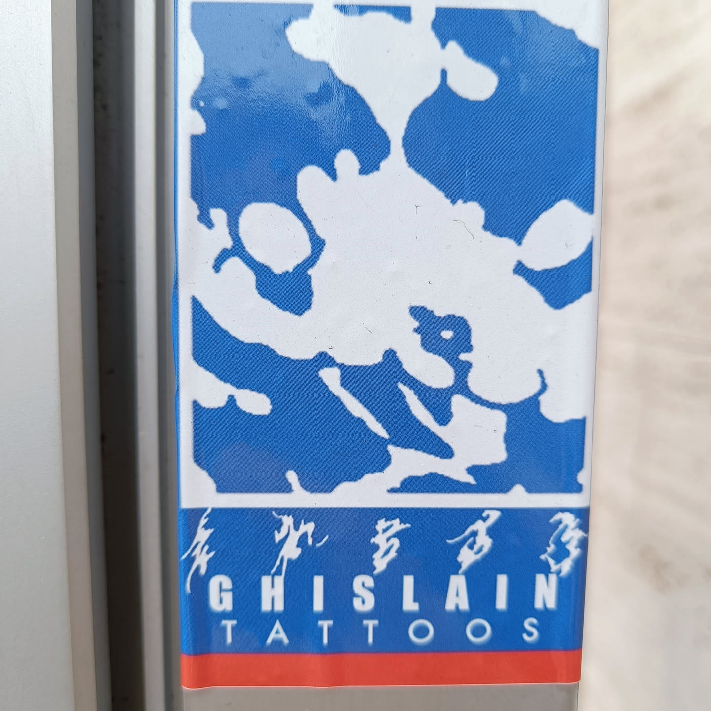 A blue and white sticker with an abstract design and the text "GHISLAIN TATTOOS" in a stylized font. There are also some Chinese characters above the name.