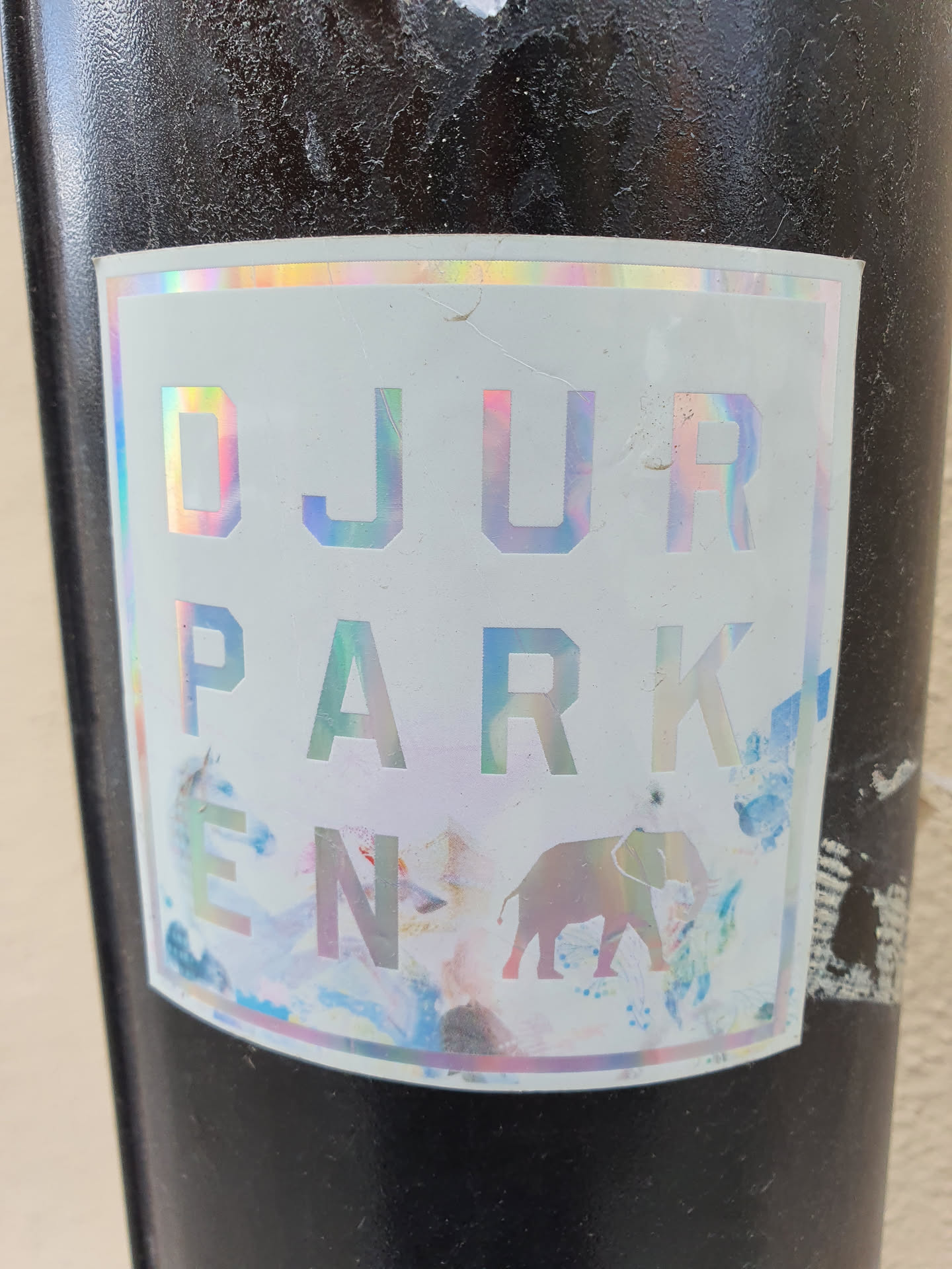 Holographic sticker with the text "DJURPARK EN" and an elephant illustration. The background features abstract watercolor-like designs in pastel colors.