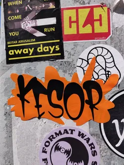 Close-up of an orange sticker with black graffiti-style lettering that says KSOP. The sticker has an irregular shape with a splatter effect. It's affixed to a wall with other stickers.