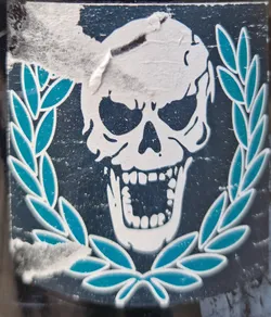 A sticker featuring a skull graphic with a laurel wreath design. The skull is white, while the wreath is teal. The background is dark. The sticker appears to be slightly damaged or worn.