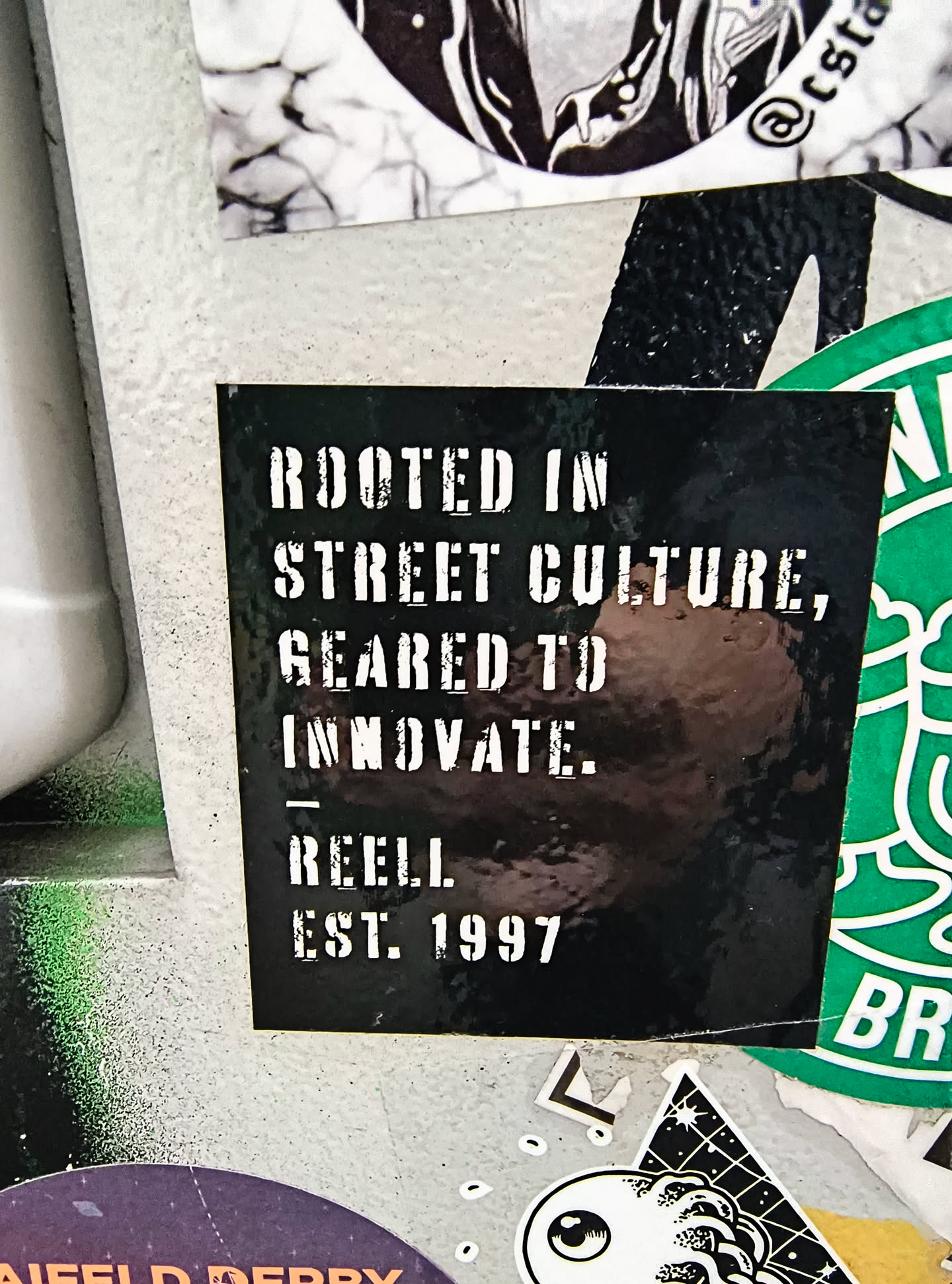 A black square sticker with white stencil-style text. The text reads: 'ROOTED IN STREET CULTURE, GEARED TO INNOVATE. REELL EST. 1997'. The sticker has a slightly glossy finish and appears to be adhered to a textured surface, possibly concrete or a similar material. There are some minor scuffs and reflections on its surface. It is layered with other stickers, with a green sticker partially visible on the right and a black and white graphic sticker above.