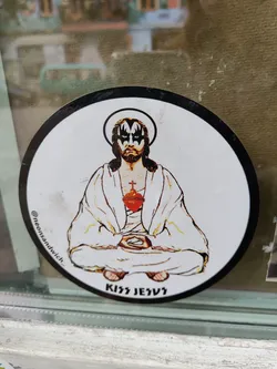 Round sticker depicting Jesus in a meditative pose with Kiss makeup. The text at the bottom reads "Kiss Jesus" and an Instagram handle is visible on the left side. 