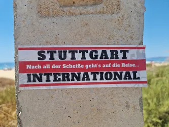 A sticker featuring the text "STUTTGART INTERNATIONAL" in bold black letters on a red and white striped background. Below the main text, there is a smaller line of German text that translates to "After all the crap, it's on to the trip...".  The sticker is affixed to a concrete post outdoors, with a beach scene visible in the background.