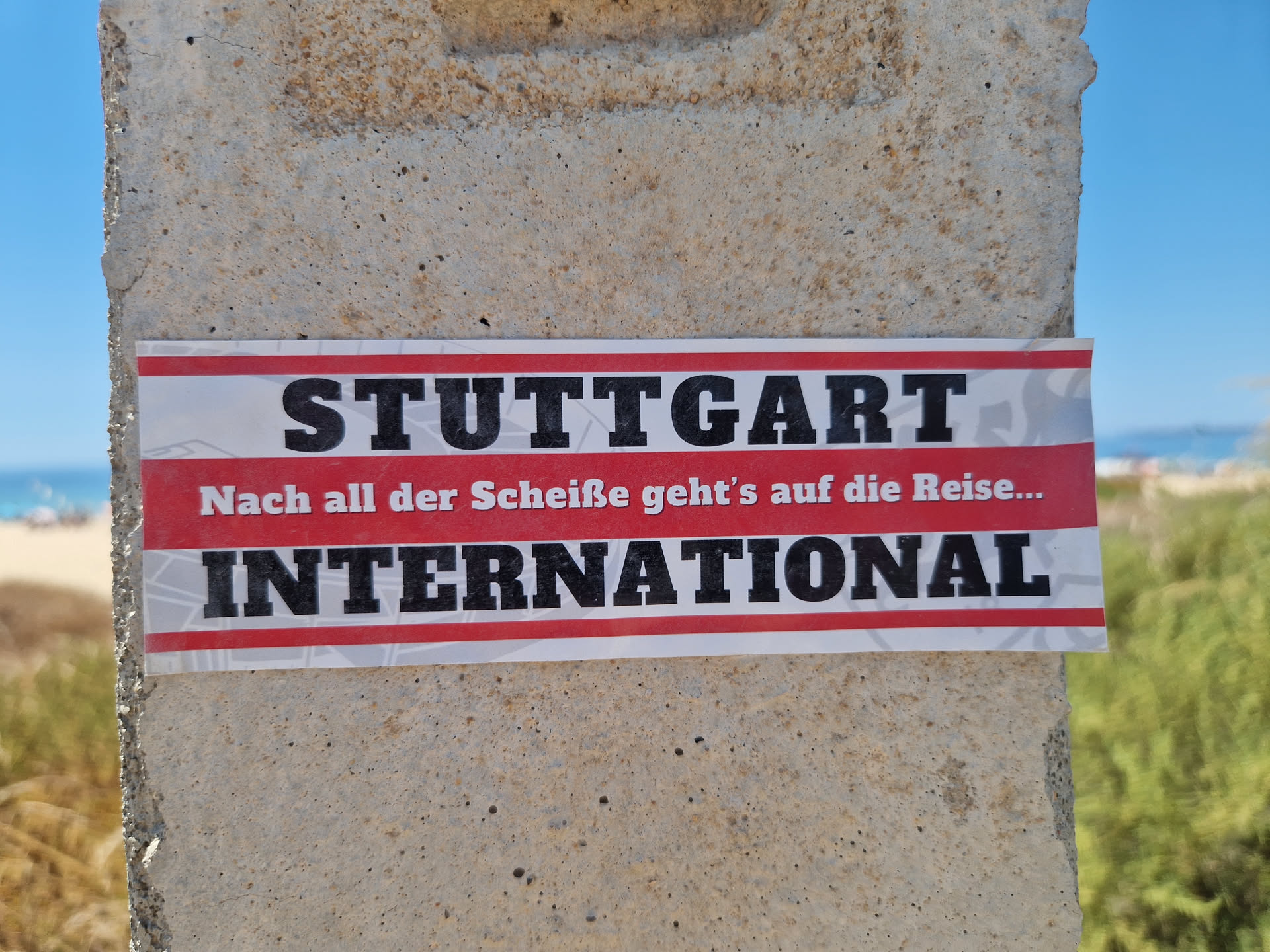 A sticker featuring the text "STUTTGART INTERNATIONAL" in bold black letters on a red and white striped background. Below the main text, there is a smaller line of German text that translates to "After all the crap, it's on to the trip...".  The sticker is affixed to a concrete post outdoors, with a beach scene visible in the background.
