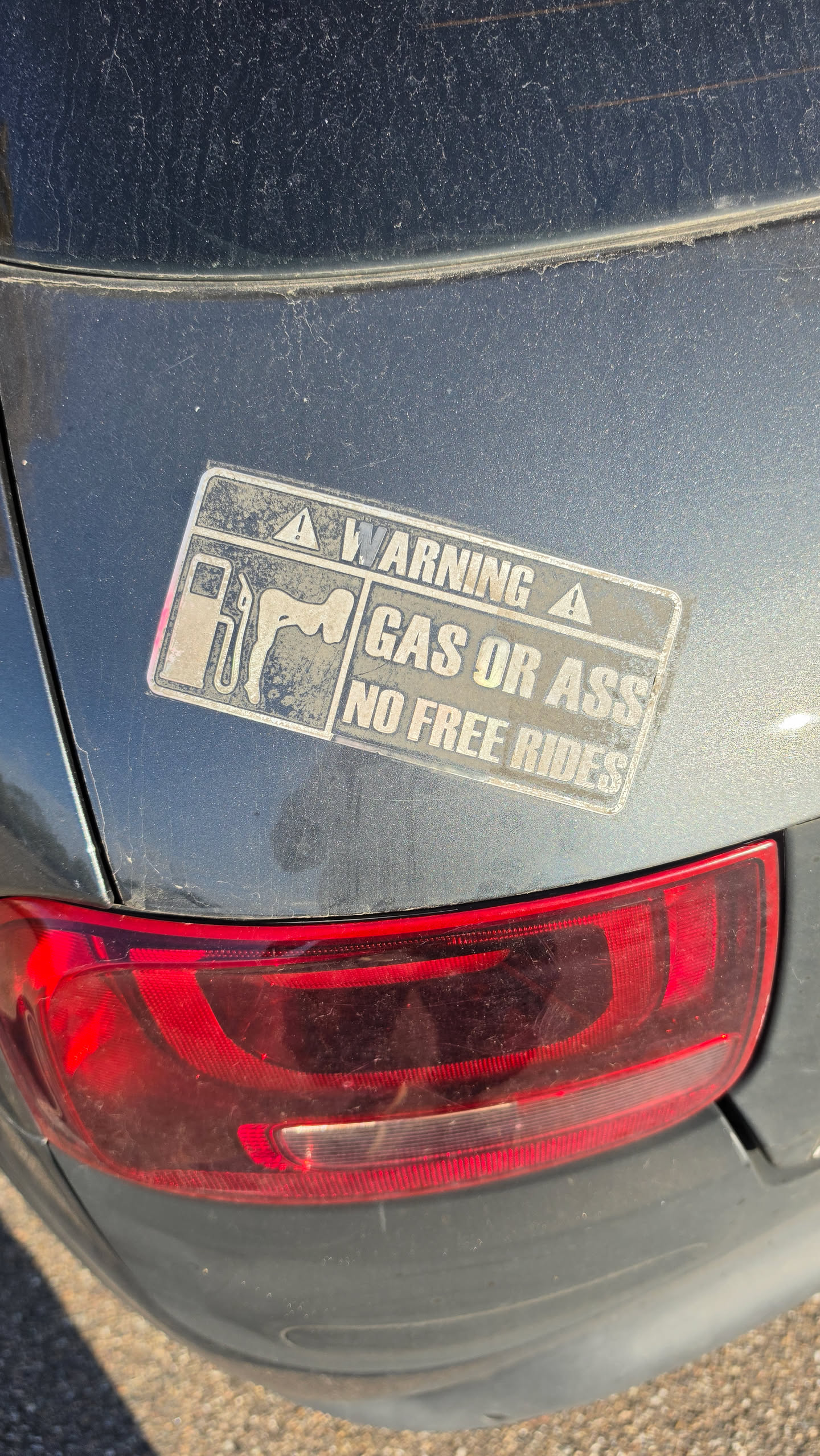 A rectangular, weathered silver bumper sticker designed to look like an official warning label. The top section features the word 'WARNING' in bold capital letters, flanked by two warning triangle icons. Below, on the left, is a graphic depicting a gas pump next to a silhouette of a person bending over. To the right of the graphic, the text reads 'GAS OR ASS' on the first line and 'NO FREE RIDES' on the second line. The sticker is applied to the dark grey metallic paint of a car, just above the red taillight, and appears dirty and worn.
