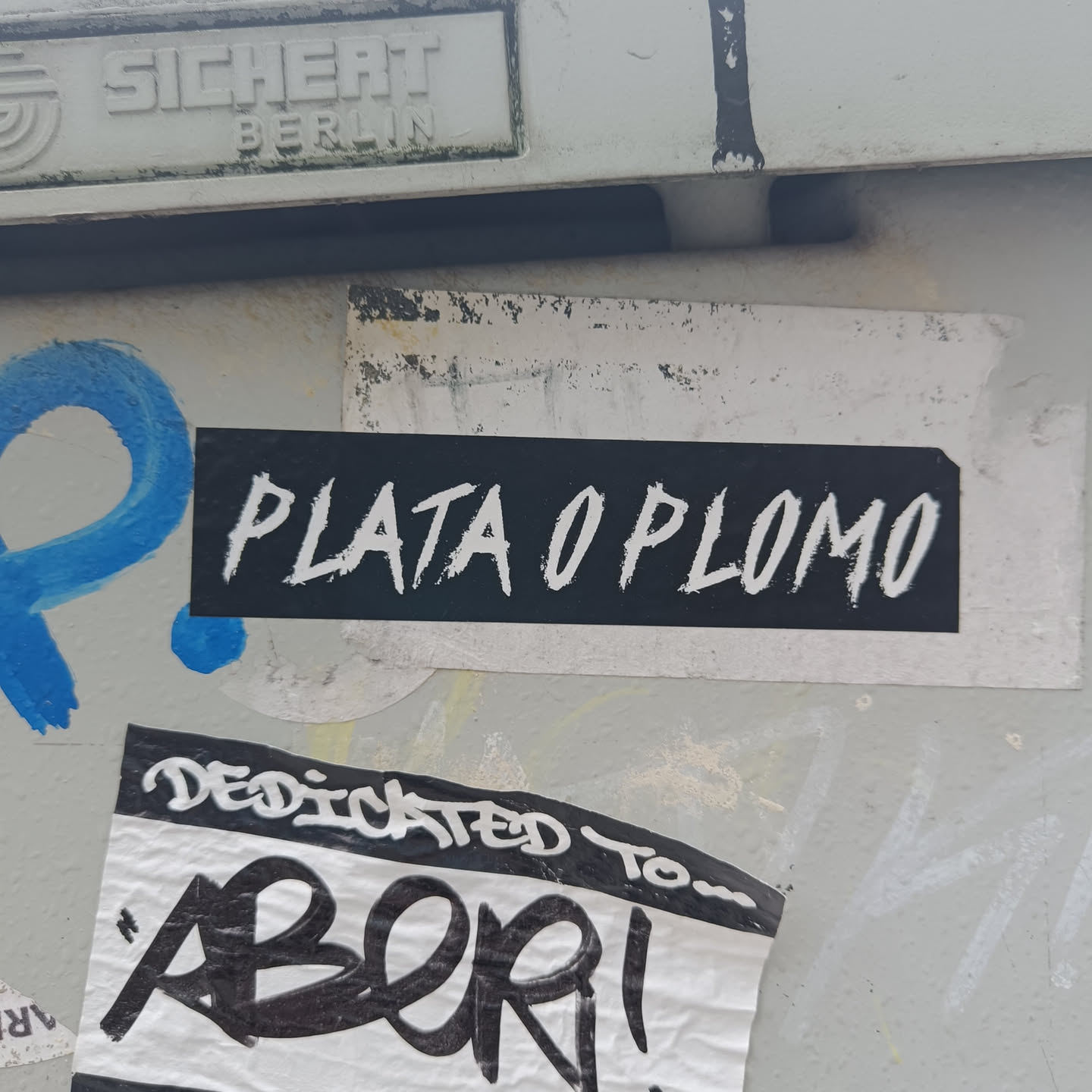 A black sticker with the text "Plata o Plomo" written in white paint. The text appears to be hand-painted, giving it a slightly rough and imperfect look.  The sticker is affixed to a wall that shows signs of other graffiti and stickers.