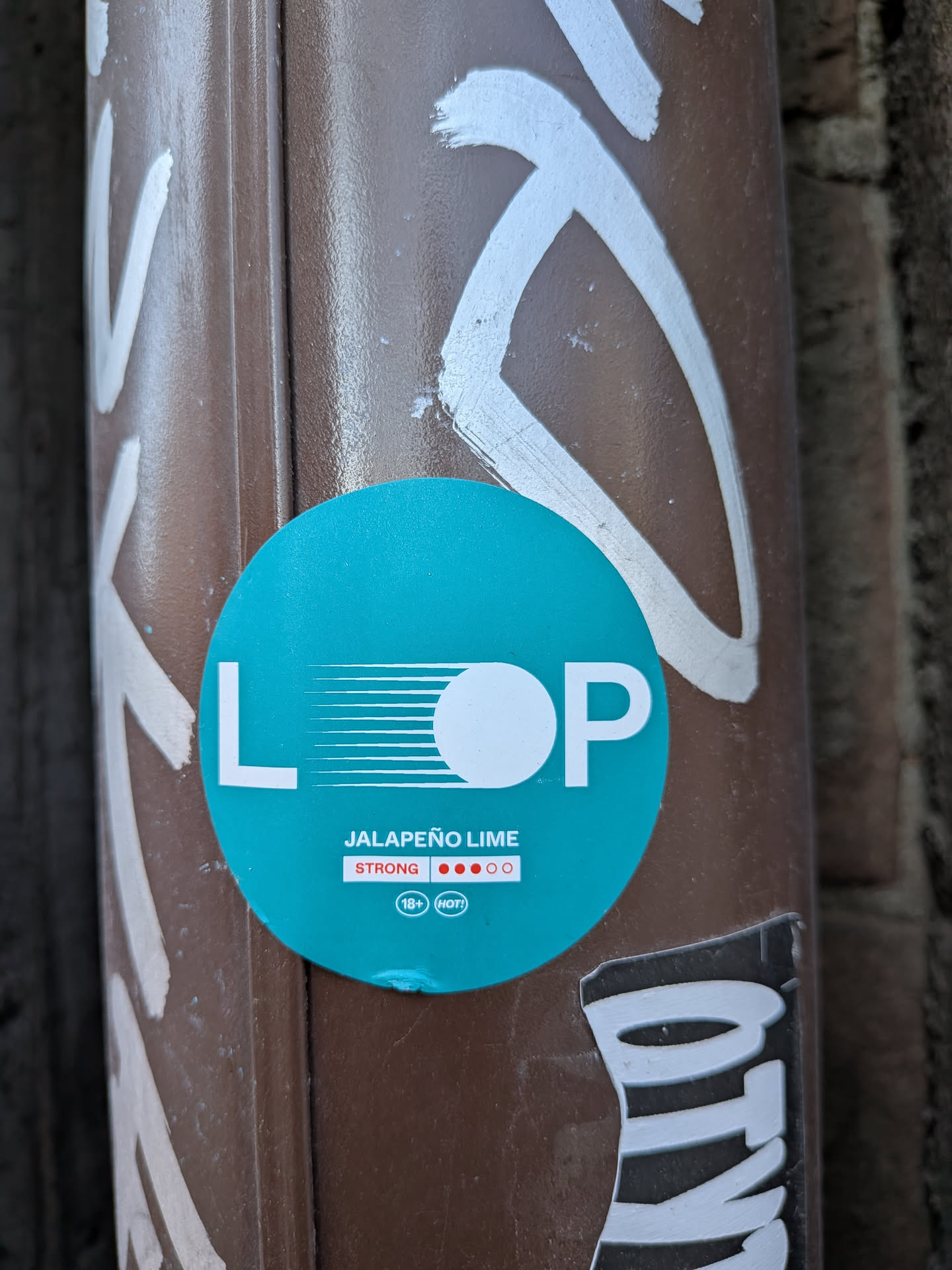 A circular teal-colored sticker prominently featuring the brand name 'L O P' in large white capital letters. The 'O' is represented by a solid white circle, with several horizontal white lines extending from the 'L' towards it, creating a dynamic visual effect. Below the main logo, the flavor 'JALAPEÑO LIME' is written in smaller white capital letters. Further down, the word 'STRONG' is displayed in white, accompanied by a visual strength indicator consisting of three solid red circles and two outlined white circles, suggesting a high potency. A small white oval contains the text '18+' and 'HOT!', indicating an adult product with a spicy profile. The sticker is affixed to a textured, dark brown pole or pipe, with remnants of white graffiti visible around its edges.