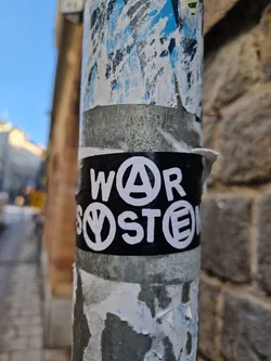 A black sticker with white lettering affixed to a metal pole. The text appears to say "WAR" with an anarchy symbol replacing the 'A', and "SYSTEM" with a peace symbol replacing the 'Y'.  The sticker is slightly worn, and there are other, older stickers visible on the pole around it. 