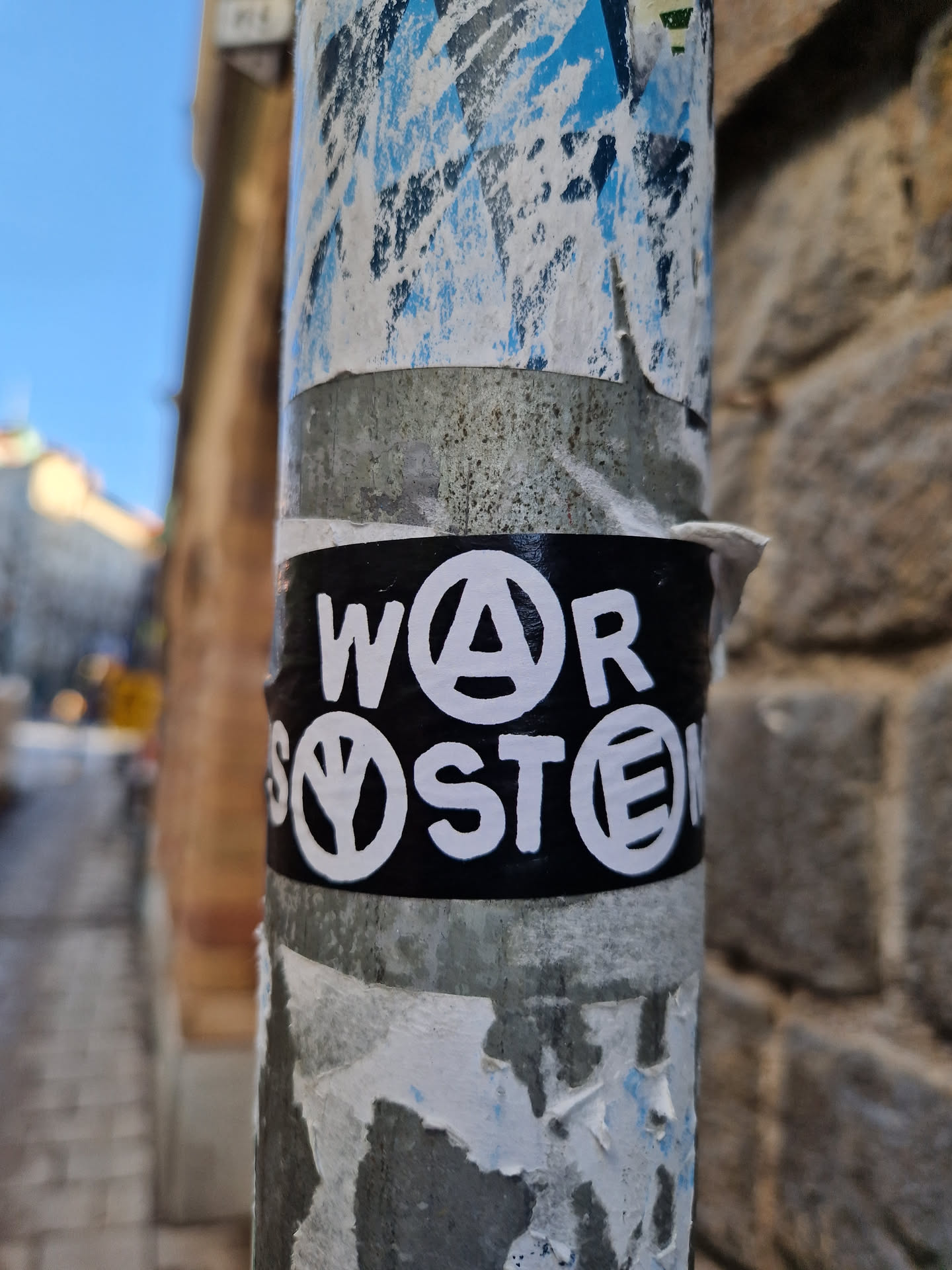 A black sticker with white lettering affixed to a metal pole. The text appears to say "WAR" with an anarchy symbol replacing the 'A', and "SYSTEM" with a peace symbol replacing the 'Y'.  The sticker is slightly worn, and there are other, older stickers visible on the pole around it. 