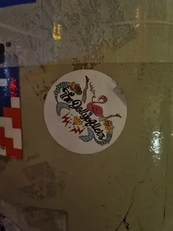 Round sticker featuring an illustration of a flamingo, a mermaid, and other quirky elements. The text 'The Dolls' is prominently displayed.