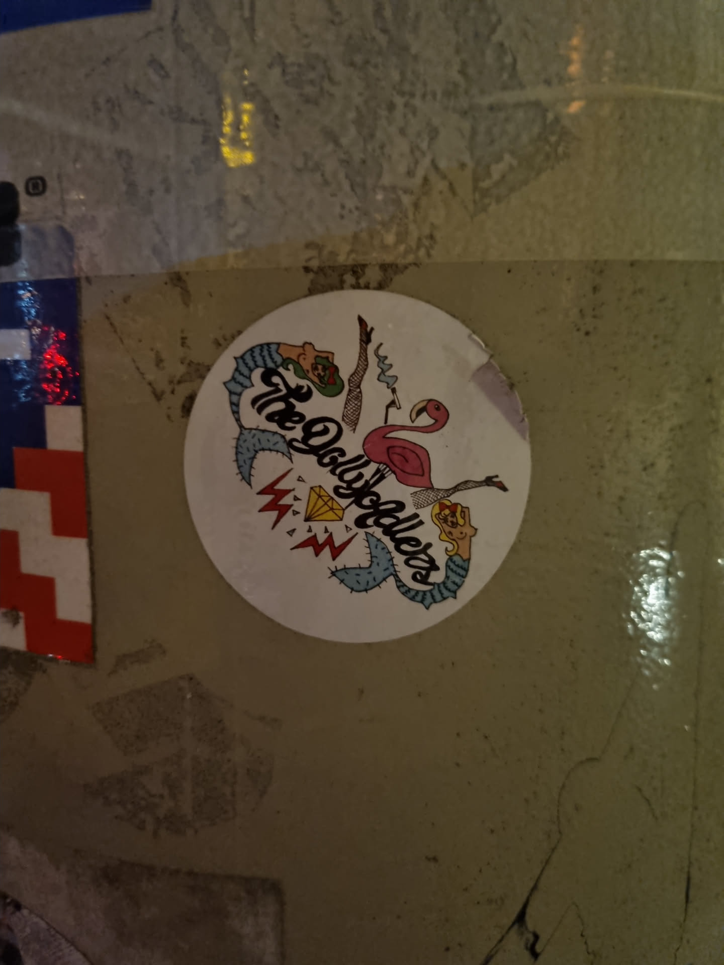 Round sticker featuring an illustration of a flamingo, a mermaid, and other quirky elements. The text 'The Dolls' is prominently displayed.