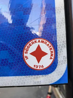 Close-up of a round sticker featuring a red star and the text 'IF Södermalmskamraterna 1974'. The sticker is affixed to a blue and white reflective surface.