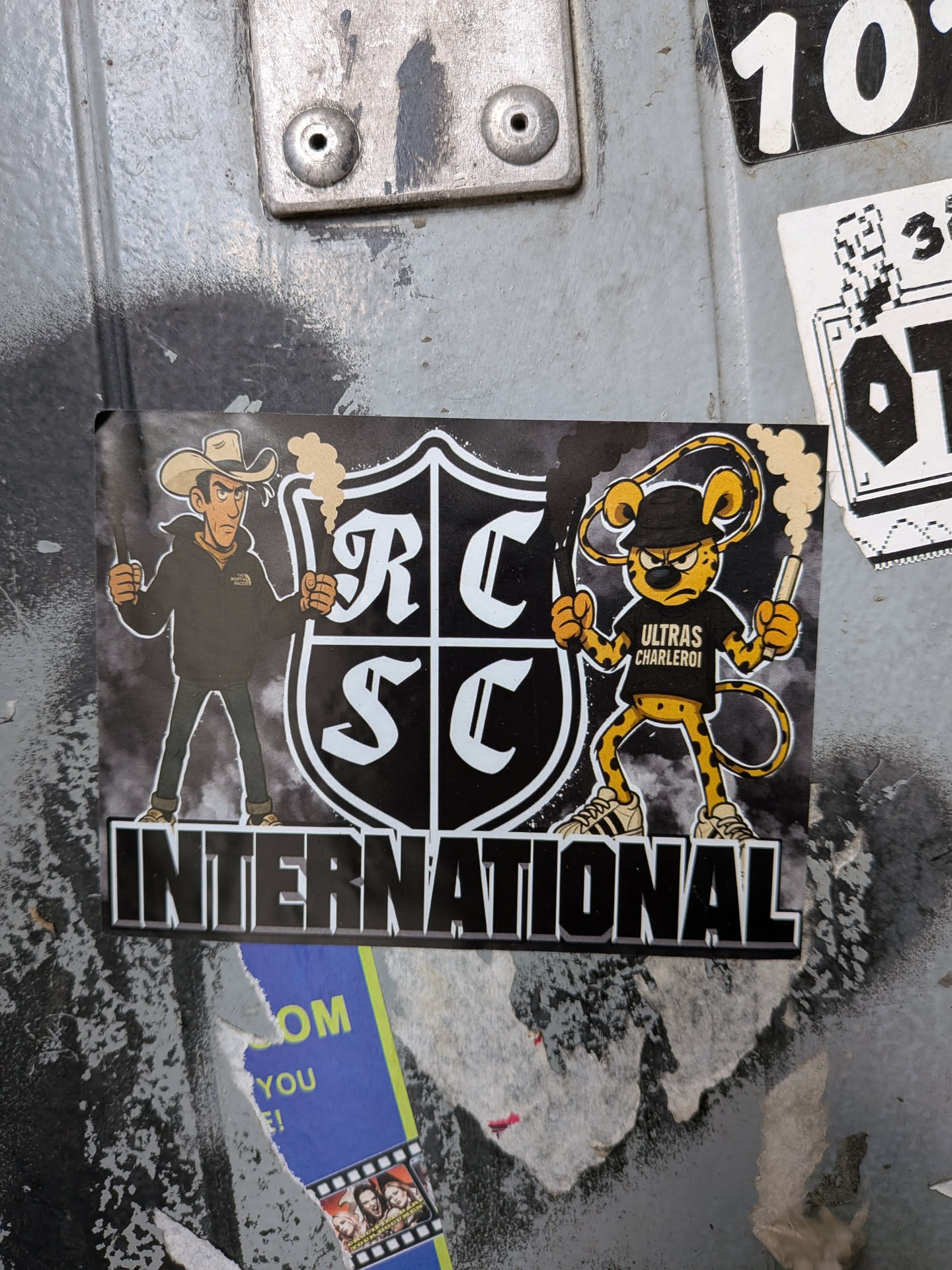 The sticker features two cartoon characters flanking a shield with 'RC SC' in large white letters. On the left is a cowboy wearing a tan hat, a black hoodie with a white logo (partially obscured), and blue jeans. He holds a lit flare in his right hand. On the right is a cheetah-like character wearing a black bandana, a black t-shirt with 'ULTRAS CHARLEROI' written in white, and white sneakers with black stripes. This character also holds a lit flare. Both characters have smoke emanating from their flares. The word 'INTERNATIONAL' is prominently displayed in large, stylized white block letters below the shield. The background of the sticker is dark with subtle textures, and the entire design appears to be printed on a rectangular sticker.