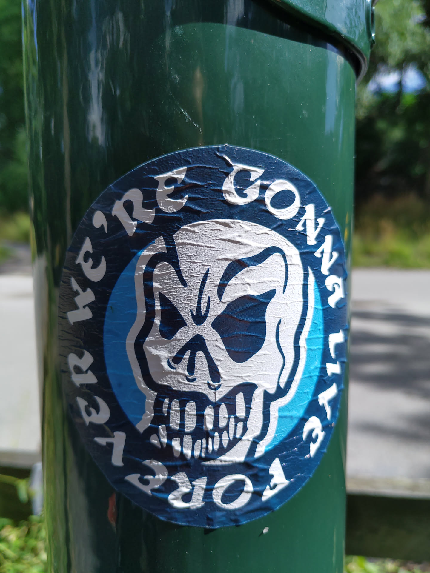 A round sticker with a skull graphic and the text "We're Gonna Die" arranged in a circular pattern. The sticker is affixed to a green pole and shows some signs of wear.