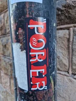 A close-up shot of a sticker on a metal pole. The sticker is predominantly black and red with the word "Porrer" written in a stylized font, accompanied by the number "031".  The sticker shows some wear and tear, indicating it's been exposed to the elements for some time. The background consists of a textured stone wall, which provides a contrast to the sticker's design.