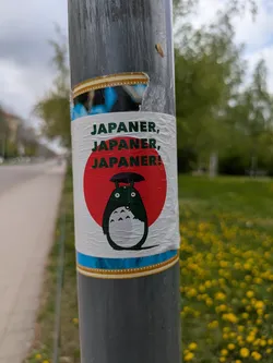 A sticker on a gray metal pole features an image of Totoro from the Studio Ghibli film "My Neighbor Totoro." The sticker is white with a red circle behind Totoro, reminiscent of the Japanese flag. Above Totoro, the text "Japaner, Japaner, Japaner!" is printed in black.