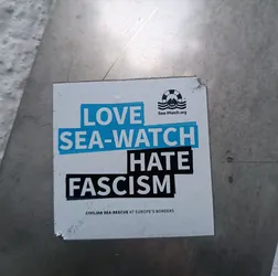 A sticker expressing support for Sea-Watch, an organization conducting civilian sea-rescue at Europe's borders, and opposition to fascism.  The sticker features the Sea-Watch logo and website address.