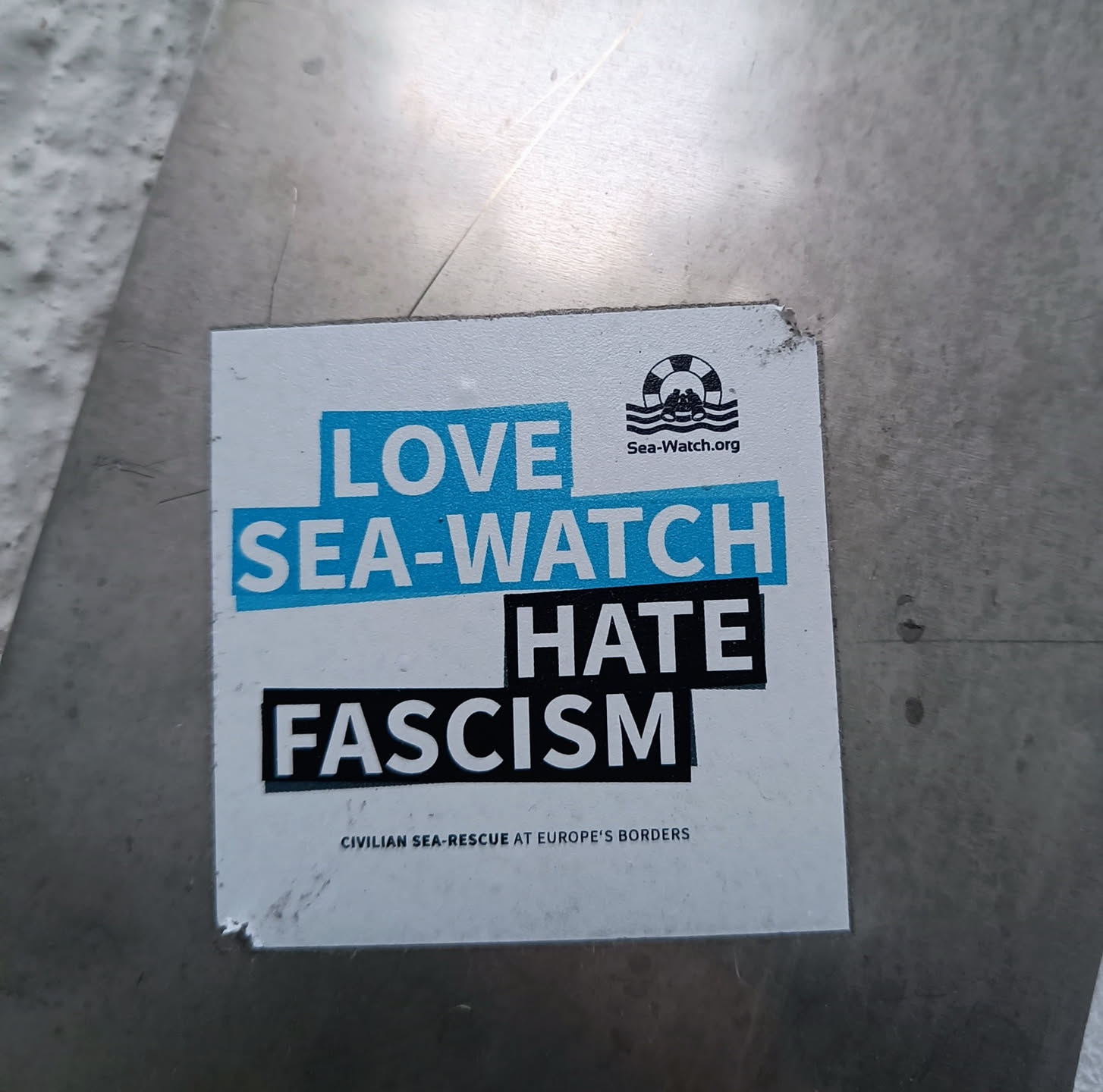 A sticker expressing support for Sea-Watch, an organization conducting civilian sea-rescue at Europe's borders, and opposition to fascism.  The sticker features the Sea-Watch logo and website address.