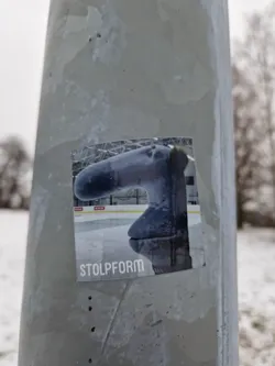 A sticker featuring a gray, stylized sculpture resembling an elephant's trunk on an ice rink. The word 'Stolpform' is printed at the bottom.  The sticker is affixed to a gray metal pole outdoors in a snowy setting.