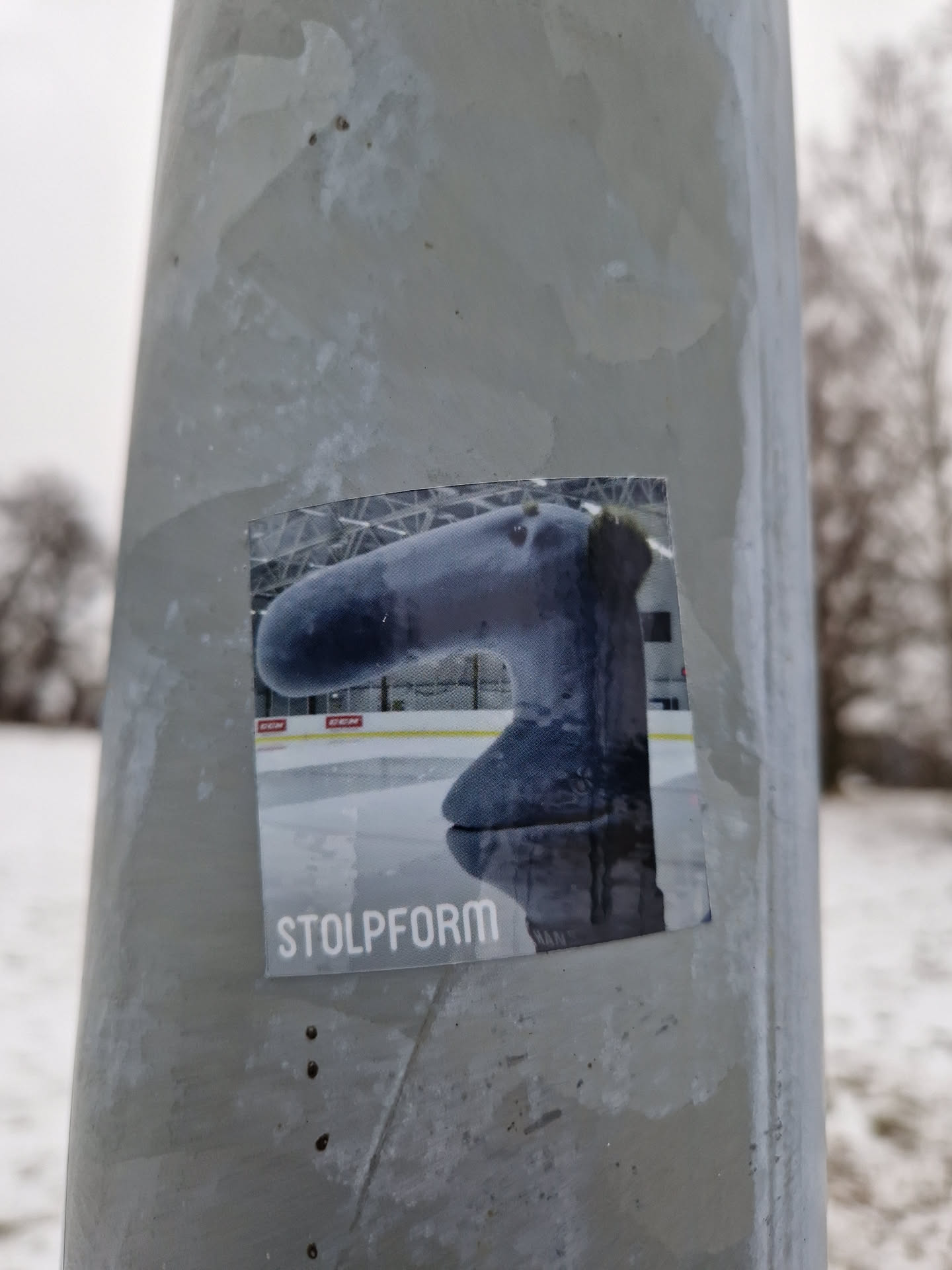 A sticker featuring a gray, stylized sculpture resembling an elephant's trunk on an ice rink. The word 'Stolpform' is printed at the bottom.  The sticker is affixed to a gray metal pole outdoors in a snowy setting.
