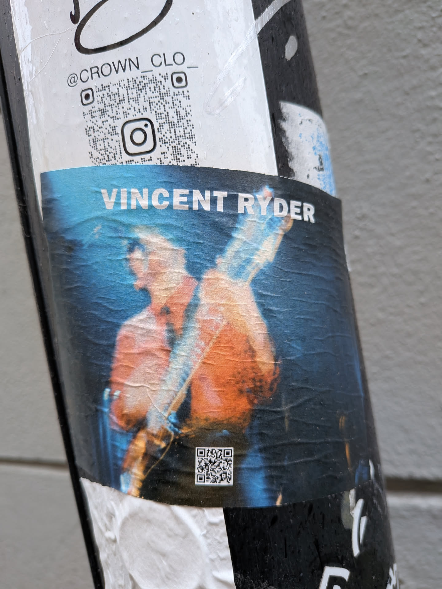 The central sticker features a dynamic, somewhat blurred image of a person, likely a musician, playing a guitar. The figure is depicted in warm, glowing orange and brown hues against a dark, moody blue background, evoking the atmosphere of a live performance under dramatic lighting. The sticker itself shows signs of wear, including wrinkles and slight tears, indicating its outdoor placement. Above the main image, the name 'VINCENT RYDER' is clearly printed in white capital letters. Below the main image, a small, black and white QR code is visible. Additionally, above this main sticker, another section on the pole displays the Instagram handle '@CROWN_CLO_' alongside a larger QR code with an Instagram logo in its center.
