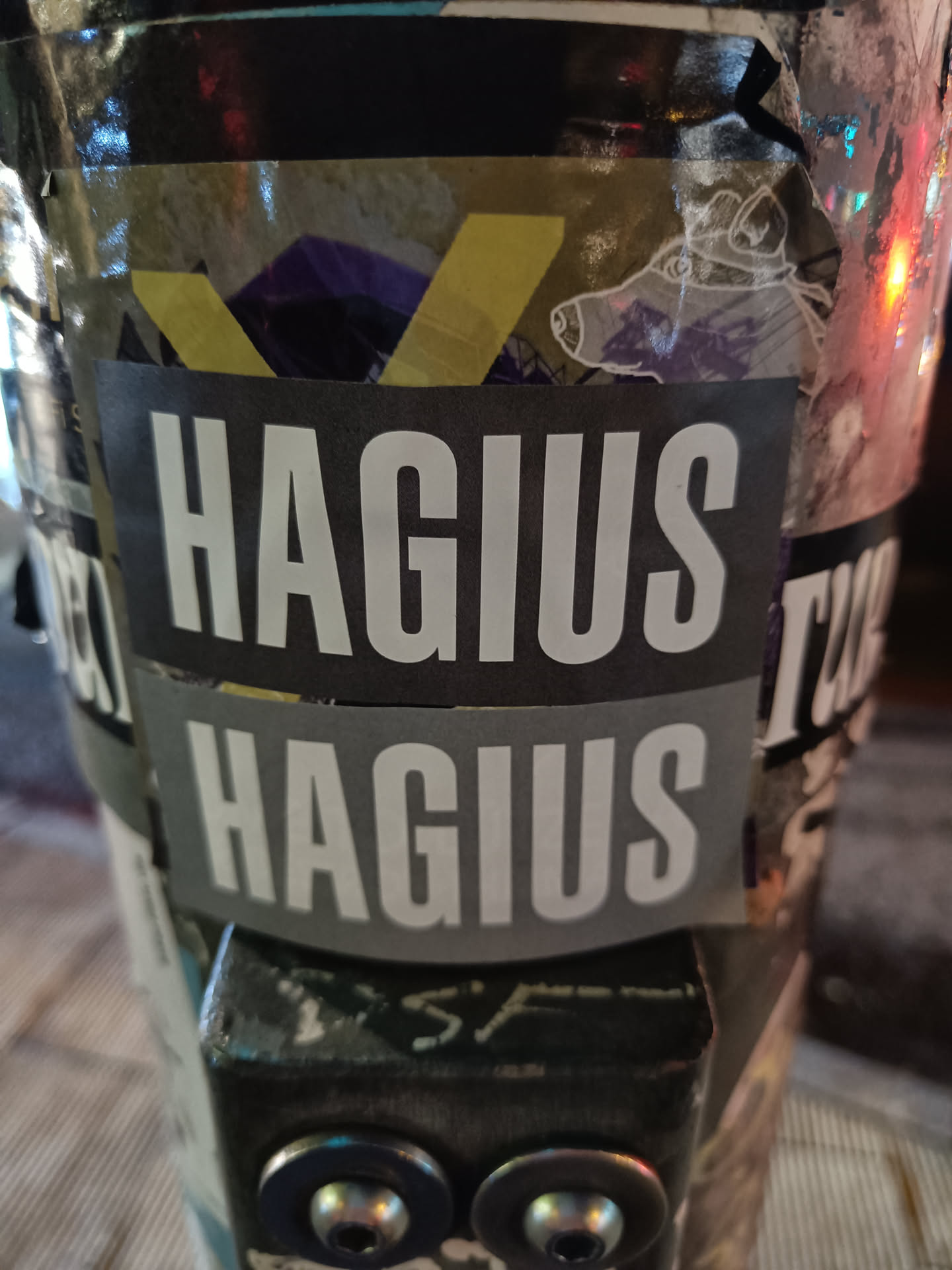 A black and grey sticker with the word "HAGIUS" repeated twice. The sticker is on a surface with other stickers.